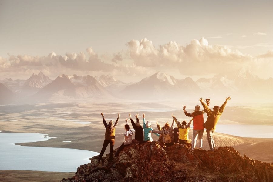 Big group of people celebrate reaching the summit of a mountain top, overlooking a beautiful valley