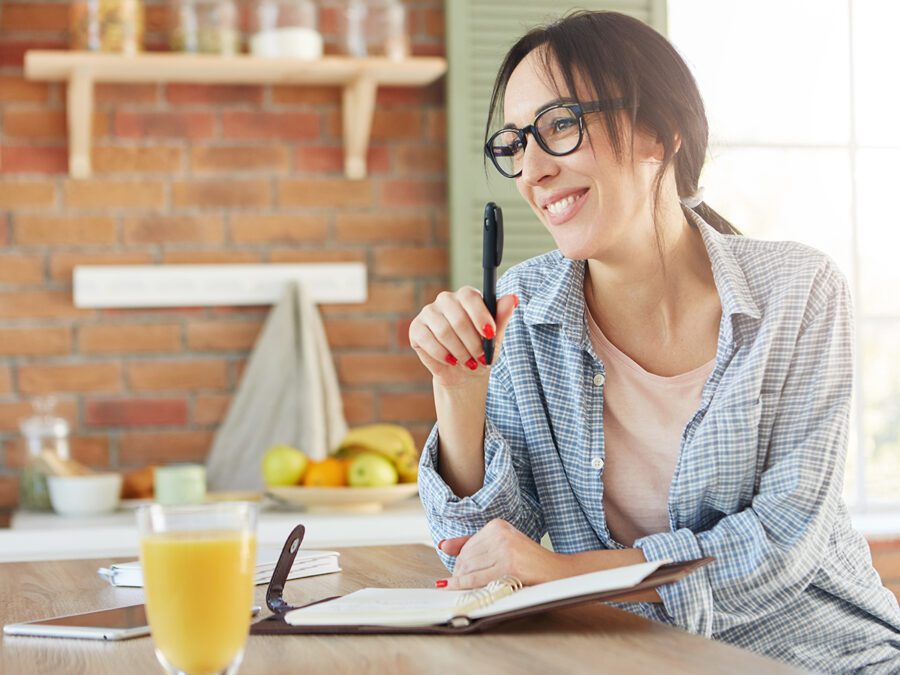 Happy woman with appealing appearance, going to organize party, makes list of invited friends, sits in kitchen, drinks orange juice, holds pen, writes in notepad salad recipe. Home atmosphere