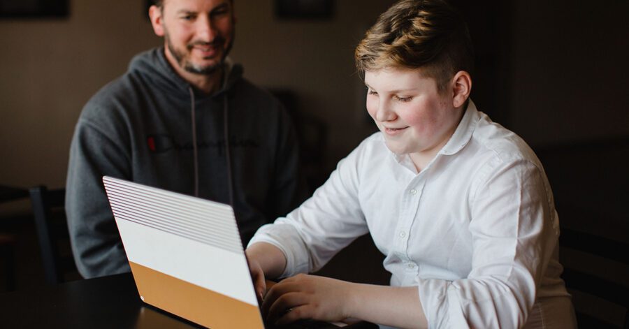 A boy in a white shirt using a laptop for homeschool while his father looks on.